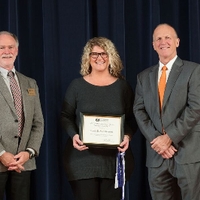Award recipient in a grey sweater dress posing for a photo on stage with Doctor Potteiger and Mark Luttenton